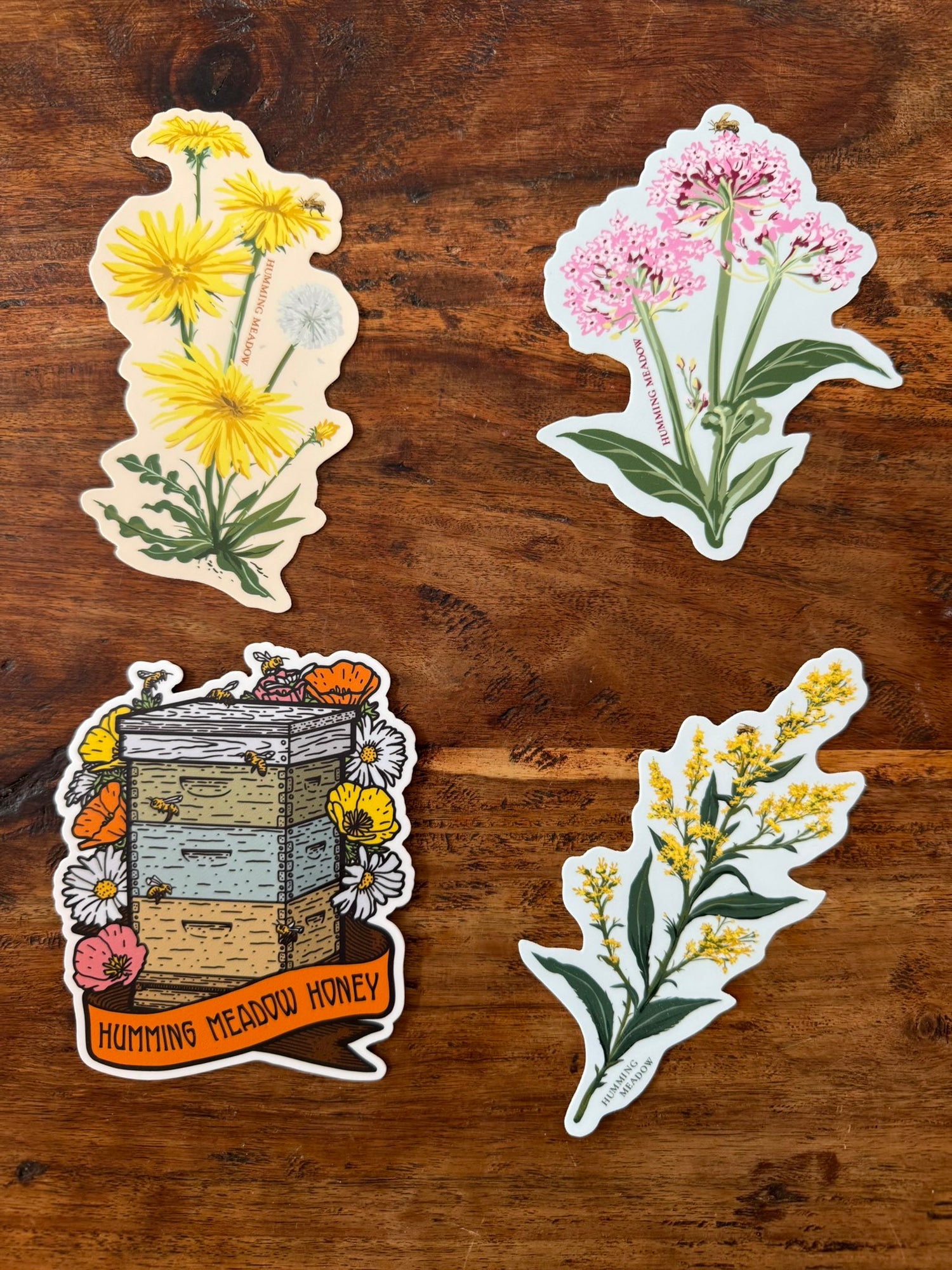 Three floral-themed stickers and one of a beehive with flowers on a wooden surface